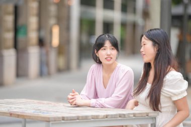 Two women sitting at a table talking. One of them is wearing a pink shirt. Scene is friendly and casual
