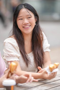 A woman is sitting at a table with two donuts in her hands. She is smiling and she is enjoying her snack