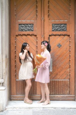 Two women standing in front of a wooden door, one holding a bouquet of flowers. Scene is lighthearted and friendly, as the two women are posing for a photo together