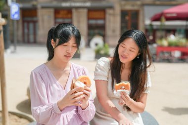 Two women are sitting on a bench eating donuts. One of the women is wearing a pink shirt