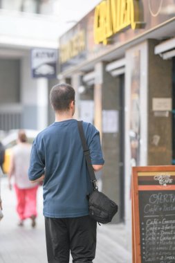 Mavi gömlekli bir adam omzunda siyah bir çantayla caddede yürüyor. Telefonuna bakıyor.