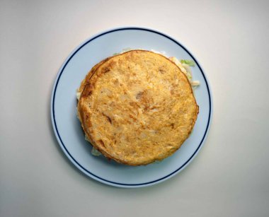 Bir restoranda servis edilen bir sebze İspanyol omletinin ağız sulandırıcı fotoğrafı, hafif bir arka planda şık beyaz ve mavi bir tabakta sunuluyor. Bu lezzetli yemek, taze sebze ve yumurtaların mükemmel bir karışımını sunuyor.