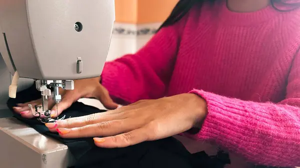 Rahat pembe bir kazak giyen genç bir kadın, siyah kumaş kırıntıları kullanarak evde dikiş dikmeyi öğreniyor. Dikiş projesine odaklanmış, öğrenme süreci ve dikiş ustalığındaki kararlılığını gösteriyor. Ev ortamı 