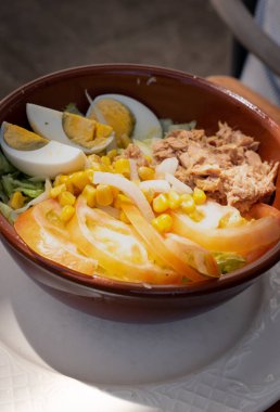 Bu resimde ton balığı, haşlanmış yumurta, tatlı mısır, dilimlenmiş domates ve taze marul karışımıyla dolu canlı bir salata kasesi yer alıyor. Malzemeler kırsal kahverengi bir kasede güzelce düzenlenmiş.