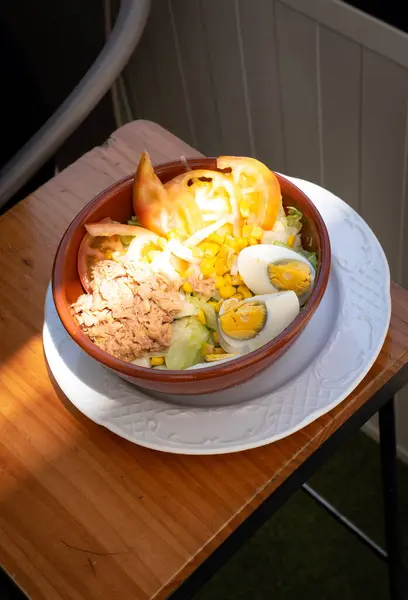 Bu resimde bir kase sağlıklı ton balığı ve taze sebzeli yumurta salatası var. Kasede dilimlenmiş haşlanmış yumurta, ton balığı parçaları, tatlı mısır ve domates dilimleri var.