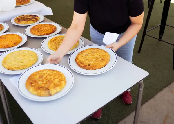Bu resim, bir açık hava etkinliğinde geleneksel İspanyol omleti tortilla espanolasının birden fazla tabağını servis eden bir kişiyi gösteriyor. Sahne, bir masaya dizilmiş çeşitli omletler yakalar.