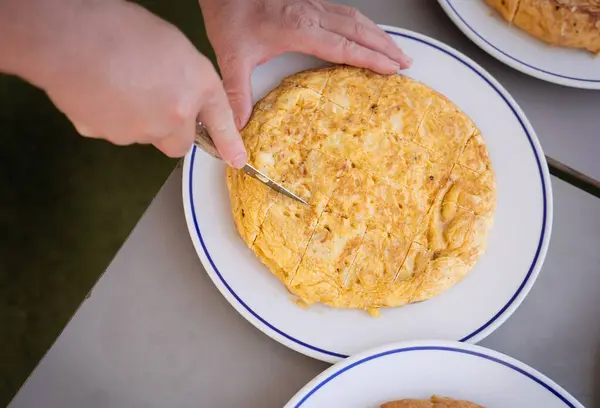 Bu üstteki görüntü, geleneksel İspanyol omlet tortilla espanolasını tahta saplı bir bıçakla dilimleme anını yakalıyor. Omlet, altın-kahverengi kabuğuyla beyaz bir tabağa konur.