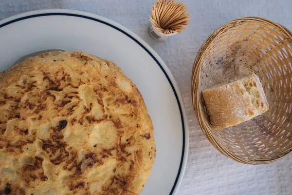 Bu yakın plan, lacivert kenarlı beyaz bir tabakta servis edilen İspanyol omletli espanola 'yı yakalar. Omletin yanında, küçük bir hasır sepette bir parça sade ekmek var.