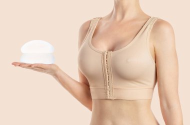 Kadın, bej arka planda sıkıştırılmış sutyen takan yuvarlak implantlar tutuyor. Estetik ameliyat.
