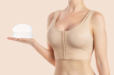 Woman holds round implants wearing compressing bra on beige background. Plastic surgery. clipart
