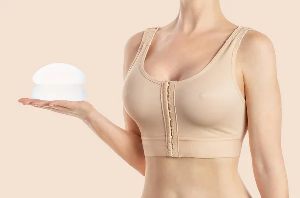 Kadın, bej arka planda sıkıştırılmış sutyen takan yuvarlak implantlar tutuyor. Estetik ameliyat.