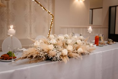 Düğün salonundaki düğün masasının süslemesi, modaya uygun boho tarzı, doğal malzemelerden yapılmış buket, kurumuş çiçekler ve otlar beyaz bir masa örtüsünün üzerinde yatıyordu.