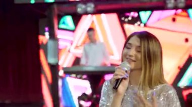 Sahnede mikrofonlu sarışın kadın şarkıcı, sahne ışıkları, interaktif ekran ve direk çerçeveye bakıyor. Burgundy elbiseli kadın ve sahte elmaslar şarkı söylüyor. Yüksek kalite 4k görüntü