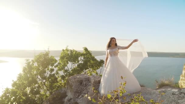 海や海の夕日に対して岩の上に立つ白いウェディングドレスとベールで若い花嫁を素晴らしい景色 風景の背景に自然の中で魅力的な若い女性 スローモーションショット — ストック動画