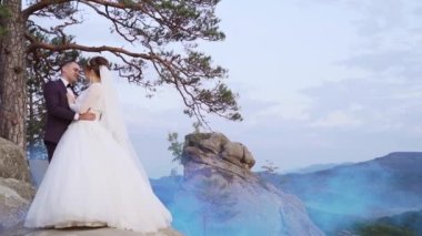 Gelin ve damat gün batımında bir uçurumda dikiliyorlar. Arka planda mavi bir sis bombası var. Yüksek kaliteli FullHD görüntüler