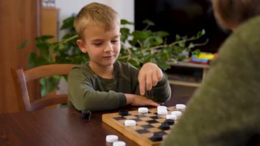 Happy little boy playing checkers with senior woman at home. Family relationship with grandmother and grandson. Grandma and male grandchild with board game. High quality 4k footage