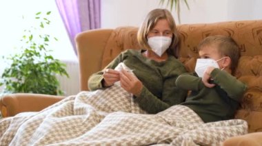 Granny and grandson in medical masks, sitting on the couch, taking temperature. High quality 4k footage