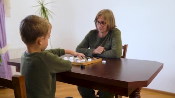 祖母と小さな孫の遊びのチェッカーがテーブルに座っている 教育委員会ゲームに従事し 複数世代の家族 子供の発達 集中力の向上 思考力 趣味の概念 — ストック動画