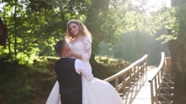 Groom spinning bride, holding her in his arms in the park near wooden bridge in slow-motion. High quality FullHD footage