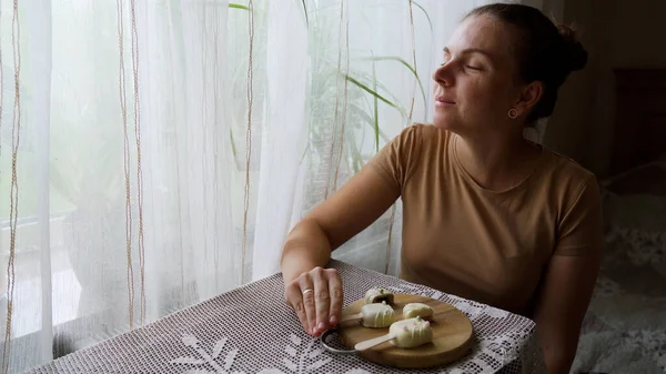 Evde tatlı yiyen bir kızın portresi. Elinde süslü pastayla tatmin olmuş bir kadın. Ev mutfağında ahşap tabaktan cheesecake yiyen çekici esmer kız. Yüksek kalite 4k görüntü