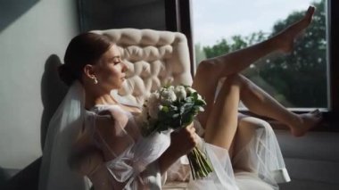 Koltuğunda dinlenen gülümseyen kadın düğün buketi ile hafifçe kendine dokunuyor. Kadın güzelliği, romantik ruh hali, evde çiçekler. Steadycam çekimi. Yüksek kaliteli FullHD görüntüler