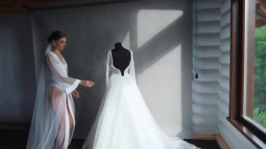 İpek Giysili Gelinlik Tutuyor ve pencerenin yanındaki güzel gelinliğine hayran. Yüksek kaliteli FullHD görüntüler