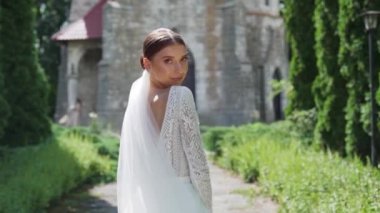 Çekici moda gelininin portresi kameraya bakıyor. Beyaz gelinlikli seksi, kendine güvenen esmer kadın kameraya poz veriyor, gülümsüyor. Kemer ve doğanın yavaş çekim arkaplanı. Dışarıda.