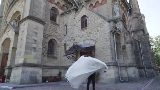 教会や城の近くの結婚式の日に彼の手に保持新郎スピニング花嫁 長い風通しの良いベールの周りに渦巻く女性は 空気中でなびく生地とエレガントなドレス 高品質のフルHd映像 — ストック動画