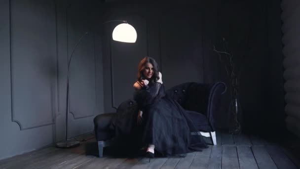 美しい黒のドレスを着て素敵な暗い髪を持つかわいいスタイリッシュな女性感情的に薄暗い光と黒の壁と部屋に大きな黒いソファの上に座って歌う 高品質4K映像 — ストック動画
