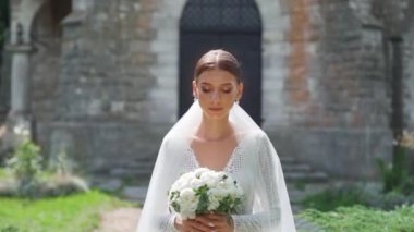 Çekici moda gelininin portresi kameraya bakıyor. Beyaz gelinlikli seksi, kendine güvenen esmer kadın kameraya poz veriyor, gülümsüyor. Kemer ve doğanın yavaş çekim arkaplanı. Dışarıda.