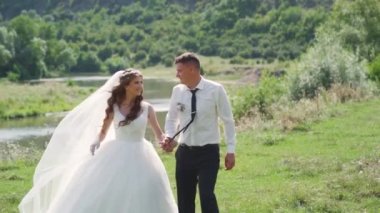 Mutlu yeni evliler, gülümsüyor ve nehir kıyısında yürüyorlar. Steadycam çekimi. Yüksek kaliteli FullHD görüntüler
