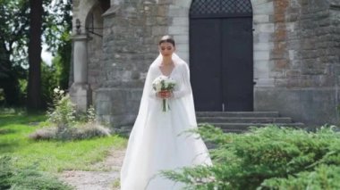 Çekici moda gelininin portresi kameraya bakıyor. Beyaz gelinlikli seksi, kendine güvenen esmer kadın kameraya poz veriyor, gülümsüyor. Kemer ve doğanın yavaş çekim arkaplanı. Dışarıda.