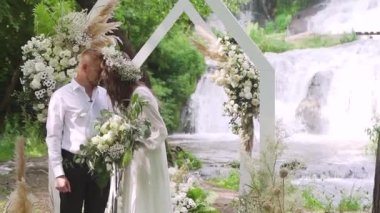 Güzel Gelin ve Damat, şelalenin yakınındaki bir açık hava düğün seremonisinde. Romantik bir çiftin evlenmesi, yüzüklerini takması ve öpüşmesi için mükemmel bir yer. Yüksek kaliteli FullHD görüntüler