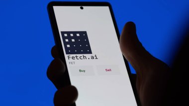 Bir yatırımcı ekranda Fetch.ai madeni parasını analiz ediyor. Bir telefon şifrenin FET 'e yatırım fiyatlarını gösteriyor