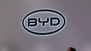 Kapalı dükkanın duvarındaki BYD logosuna odaklan. Elektronik bir galeride BYD markası. Önplanda bulanık bir yolcunun parçası.