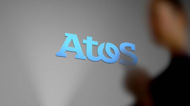 Atos 'un logosu gri bir duvarda, ön planda bulanık bir yolcu..