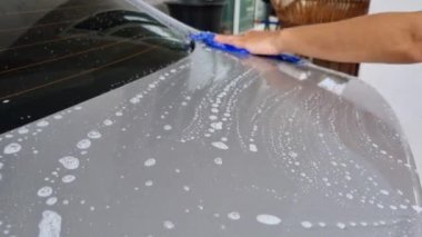 Asyalı bir adam su hortumu kullanarak kendi arabasını yıkıyor. Sedanın arkasında sabunlu köpükle toz bezi ve leke kullanarak.