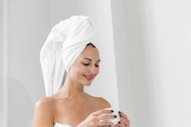 Güzellik düşkünü bir kadın sağlıklı, pürüzsüz bir vücudu olan bir havlu giyiyor, büyük pencereli bir banyoda beyaz bir küvette kahve içiyor..