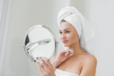 A beauty-loving woman wearing a towel with a healthy, smooth body looking at a round mirror in a white bathtub in a bathroom.	