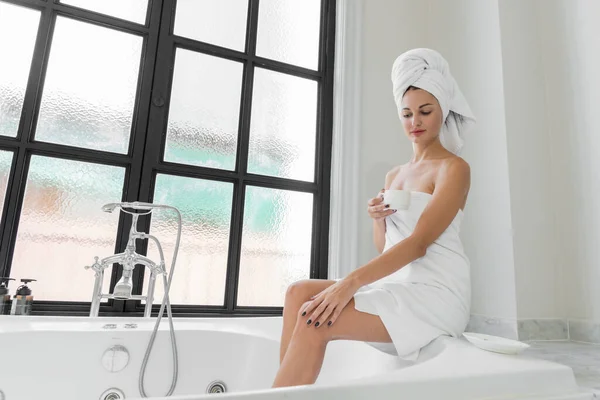 stock image A beauty-loving woman wearing a towel with a healthy, smooth body, drinking coffee in a white bathtub in a bathroom with a large window.