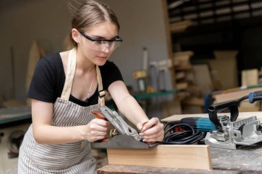 Mobilya fabrikasında mobilya aletleri kullanan bir kadın marangozun portresi. Modern aletlerle mobilya yapmak için kullanılan ahşap bir planör kullanıyor.