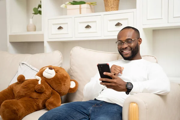 Afrikalı adam akıllı telefonuyla oynuyor. Evdeki kanepede dinleniyor.