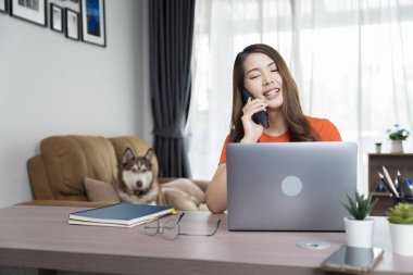 Kıvırcık saçlı Asyalı kadın, bilgisayar ve cep telefonuyla iletişim kanalları, telefon, internet üzerinden müşterileri veya iş arkadaşlarıyla sohbet ediyor.
