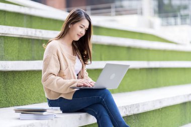 Asyalı kız öğrenci üniversite alanında sınavlara hazırlanıyor. İnternete bağlı cep telefonu ya da dizüstü bilgisayarla iletişim kurun. bir çok bilgi kaynağıyla