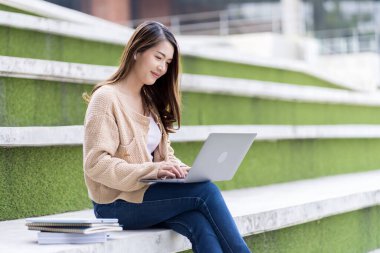 Asyalı kız öğrenci üniversite alanında sınavlara hazırlanıyor. İnternete bağlı cep telefonu ya da dizüstü bilgisayarla iletişim kurun. bir çok bilgi kaynağıyla