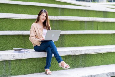 Asyalı kız öğrenci üniversite alanında sınavlara hazırlanıyor. İnternete bağlı cep telefonu ya da dizüstü bilgisayarla iletişim kurun. bir çok bilgi kaynağıyla