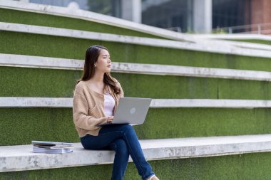 Asyalı kız öğrenci üniversite alanında sınavlara hazırlanıyor. İnternete bağlı cep telefonu ya da dizüstü bilgisayarla iletişim kurun. bir çok bilgi kaynağıyla