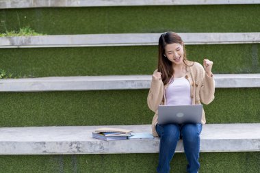 Asyalı kız öğrenci üniversite alanında sınavlara hazırlanıyor. İnternete bağlı cep telefonu ya da dizüstü bilgisayarla iletişim kurun. bir çok bilgi kaynağıyla