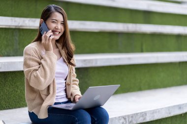 Asyalı kız öğrenci üniversite alanında sınavlara hazırlanıyor. İnternete bağlı cep telefonu ya da dizüstü bilgisayarla iletişim kurun. bir çok bilgi kaynağıyla