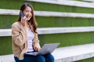 Asyalı kız öğrenci üniversite alanında sınavlara hazırlanıyor. İnternete bağlı cep telefonu ya da dizüstü bilgisayarla iletişim kurun. bir çok bilgi kaynağıyla